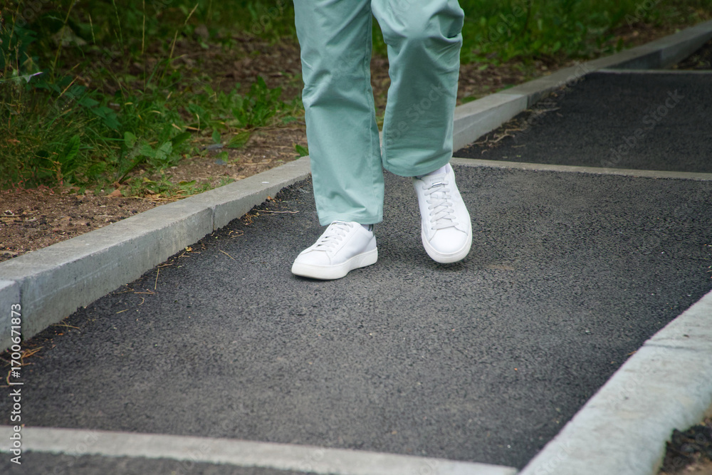 Fototapeta premium Woman’s legs in mint pants walking on asphalt steps in park, wearing white sneakers. Outdoor lifestyle and casual fashion detail. Photo