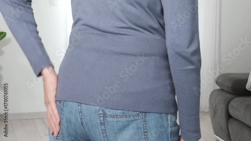 Back pain. Middle aged woman holding her lower back and massaging it with her hands at home. Close-up, rear view, side view. Lumbago, sciatica, lower back pain, lumbago in the back, hernia, protrusion