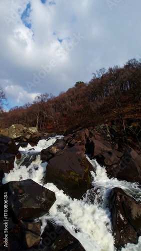 Waterfall cascading over dark rocks, with a backdrop of trees and a partly cloudy sky