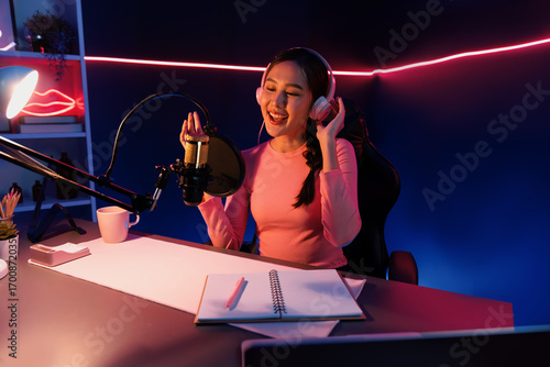 Foto Host channel of young beautiful Asian broadcaster listening the music on social media with listeners, wearing pastel headphones, using microphone for singing at neon light indoor studio