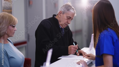 An elderly man signs a form at a medical reception desk while his companion waits.