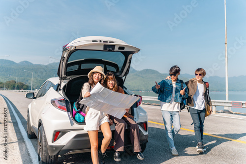 Happy Young Asian man and woman friends road trip travel countryside on summer holiday vacation. Generation z people enjoy outdoor active lifestyle sit on EV car trunk and looking direction on a map.