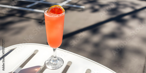 Fototapeta Naklejka Na Ścianę i Meble -  Tall glass of vibrant pink grapefruit cocktail on a white chair with sunlight and shadows on a parking area background