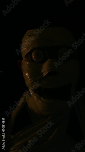Vertical video of portrait of man in creepy mummy wrapped custom with bandages wearing his mask and moving his face to look funny and creepy, dark lighting