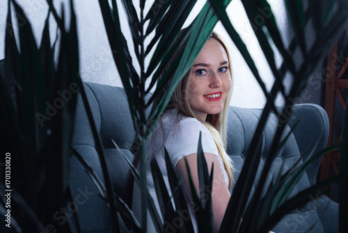 In a cozy living room, a young woman looks out from behind the leaves of plants
