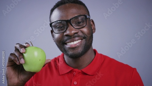 Smiling Afro-American man in spectacles holds green apple in front of the camera and smiles, displaying healthy lifestyle on grey background. Red shirt, positive emotions, diet. Male portrait, close