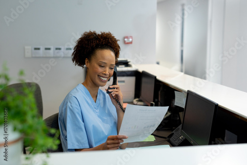 Hospital receptionist in scrubs smiling while talking on phone and holding document
