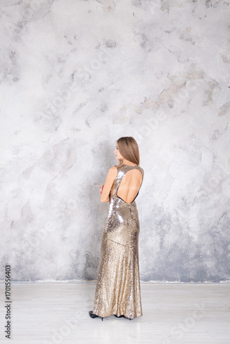 Woman standing in a long gold sequined dress against a textured gray wall, looking over her shoulder