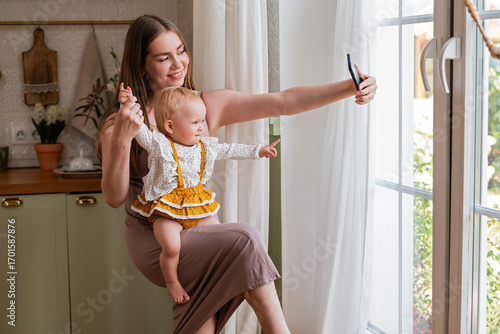 Young mother taking selfie with baby in her arms in kitchen.