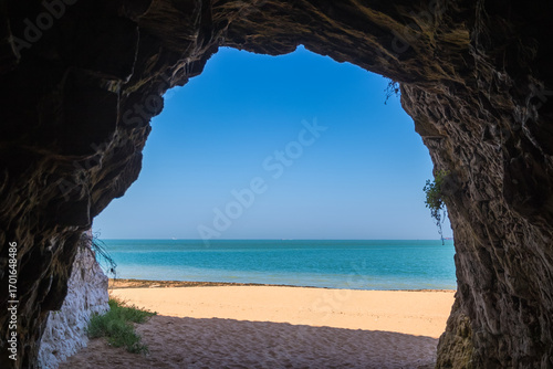 A scenic view from inside a seaside cave at Kingsgate Bay in Broadstairs, England, looking out onto a sunlit sandy beach and the calm turquoise sea under a clear blue sky