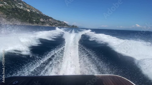 Picturesque foam wake on crystal clear blue water surface from riding boat on high speed along Amalfi coast, panoramic view of seascape and rock hills with scattered houses, recreation during summer