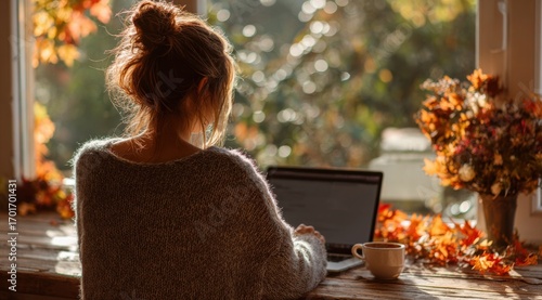 A Cozy Autumn Scene Featuring a Young Woman Working on a Laptop by a Sunlit Window Surrounded by Fall Leaves and a Warm Cup of Coffee