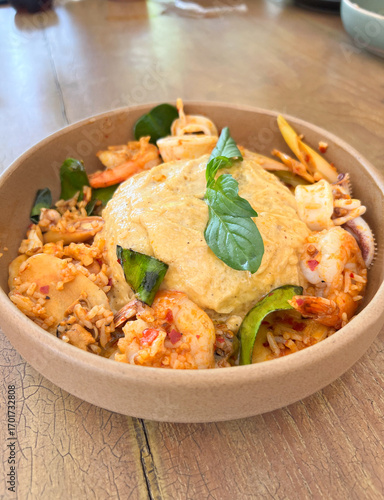 focus on fresh basil leaves placed on top of thai local food ,spicy seafood fried rice mixed with shrimp, squid, vegetables, and chili topped with fluffy eggs omelet served in a brown ceramic plate. 