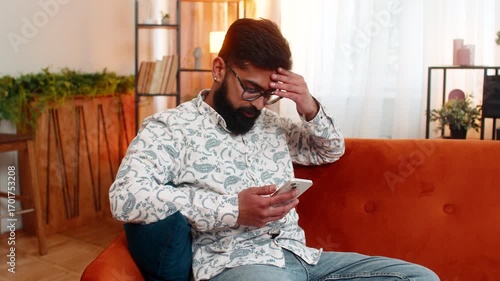 Indian man at home receives bad news on smartphone while sitting on sofa after losing online game. Arabian guy looks upset, shakes head, showing frustration and disappointment in stressful moment.