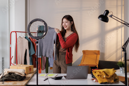 Young Asia lady seller look at phone camera live stream use laptop answer follower comment introduce sweater new collection new arrival on her chanel at studio. Woman fashion online sme shop concept.