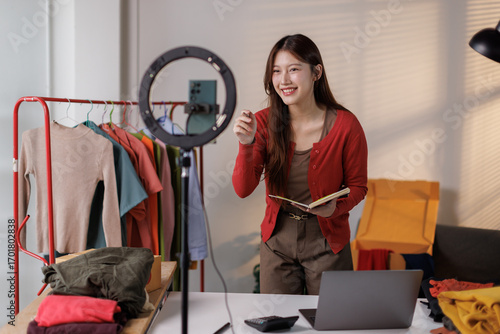 Young Asia lady seller look at phone camera live stream use laptop answer follower comment introduce sweater new collection new arrival on her chanel at studio. Woman fashion online sme shop concept.