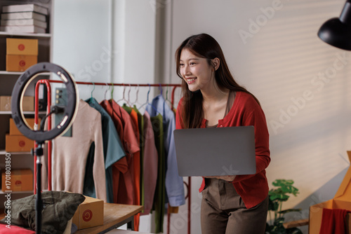 Young Asia lady seller look at phone camera live stream use laptop answer follower comment introduce sweater new collection new arrival on her chanel at studio. Woman fashion online sme shop concept.