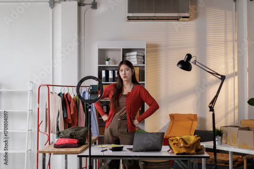 Young Asia lady seller look at phone camera live stream use laptop answer follower comment introduce sweater new collection new arrival on her chanel at studio. Woman fashion online sme shop concept.