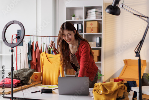 Young Asia lady seller look at phone camera live stream use laptop answer follower comment introduce sweater new collection new arrival on her chanel at studio. Woman fashion online sme shop concept.