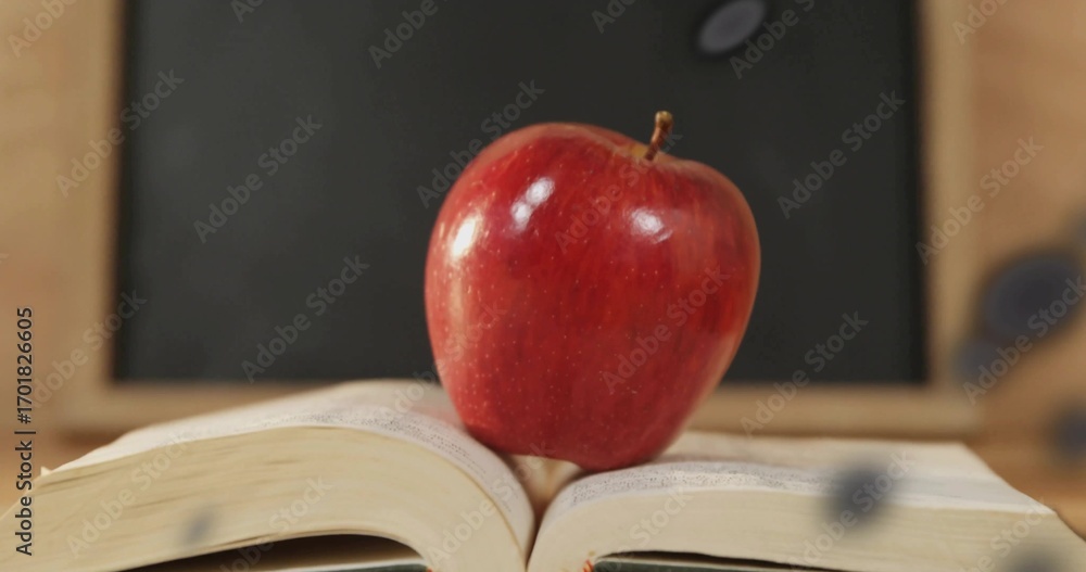 Naklejka premium Sitting red apple balancing on open book on desk in classroom, with chalkboard and chalk dust