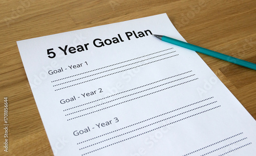 Close up of a paper sheet that says '5 Year Goal Plan' with a pencil pointing at the headline, isolated on wooden background