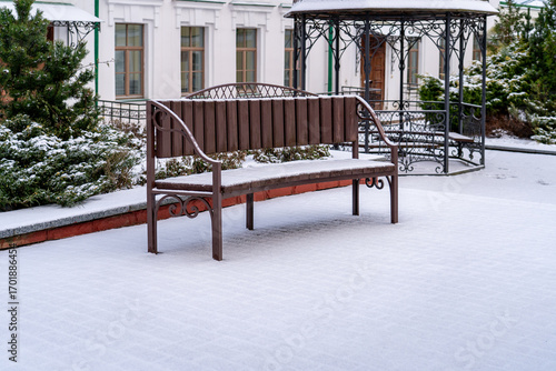 Charming brown park bench, lightly dusted with fresh snow, rests on paved winter ground with decorative gazebo and white building in serene background.
