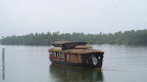 Traditional houseboat with cane bamboo roof floating away in the rain in a glassy smooth backwater in Bekal Kovalam kerela with trees in the distance showing popular tourist activity
