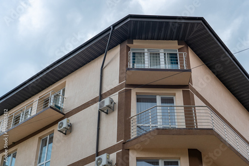 Modern multi-story building with beige and brown facade, contemporary balconies, numerous air conditioning units, and dark angled roof against cloudy sky