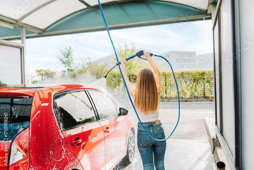 Young woman washing red car with pressure hose outdoors