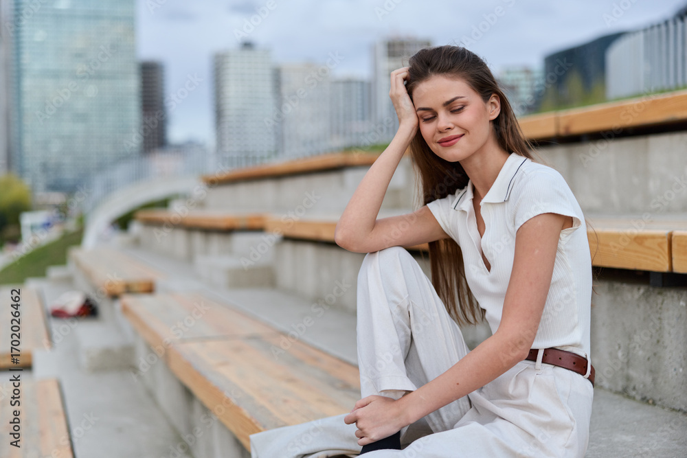 Fototapeta premium Young woman sitting relaxed outdoors in casual white outfit with modern urban background, smiling peacefully, lifestyle and fashion concept