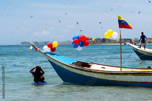 Dos niños están bañándose en una playa al lado de una lancha con globos y una bandera de Venezuela en una playa de la Isla de Margarita.