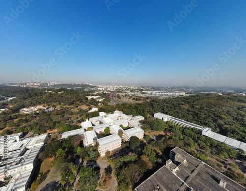 Drone view of the UFMG campus in Belo Horizonte, Brazil, with the iconic Mineirão and Mineirinho stadiums visible, surrounded by greenery and the city skyline stretching into the horizon