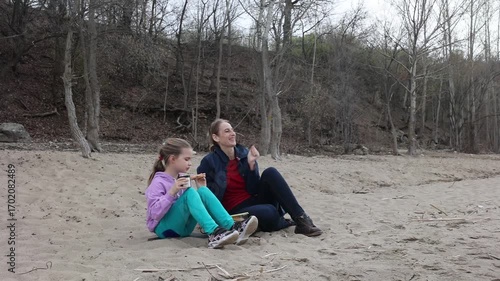 family mom and daughter is eight years old drinking tea and eating sandwiches sitting on the sand near the river on a spring day.  Family leisure with kids at spring.