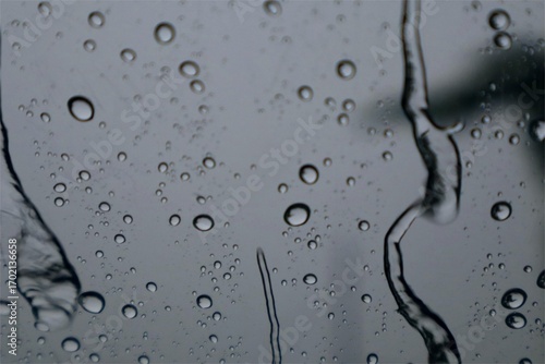 Close-up of raindrops on a glass window with a blurred background, featuring an abstract water texture and natural rainfall pattern that creates a moody and artistic atmosphere.