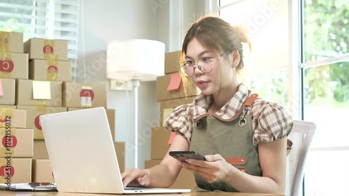 Young Asian female entrepreneur manages online business orders from home using smartphone, laptop and e-commerce work package.