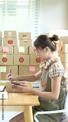 Young Asian female entrepreneur manages online business orders from home using smartphone, laptop and e-commerce work package.
