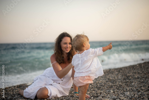 Mother holding baby at the seaside during summer sunset. Happy family moment on the beach with ocean view. Concept of motherhood, childhood, love, and vacation lifestyle outdoors.