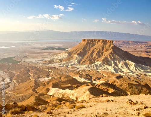A high-angle view of a dramatic desert landscape, featuring a mesa-like mountain, arid valleys, and a vast, pale-toned body of water in the distance.