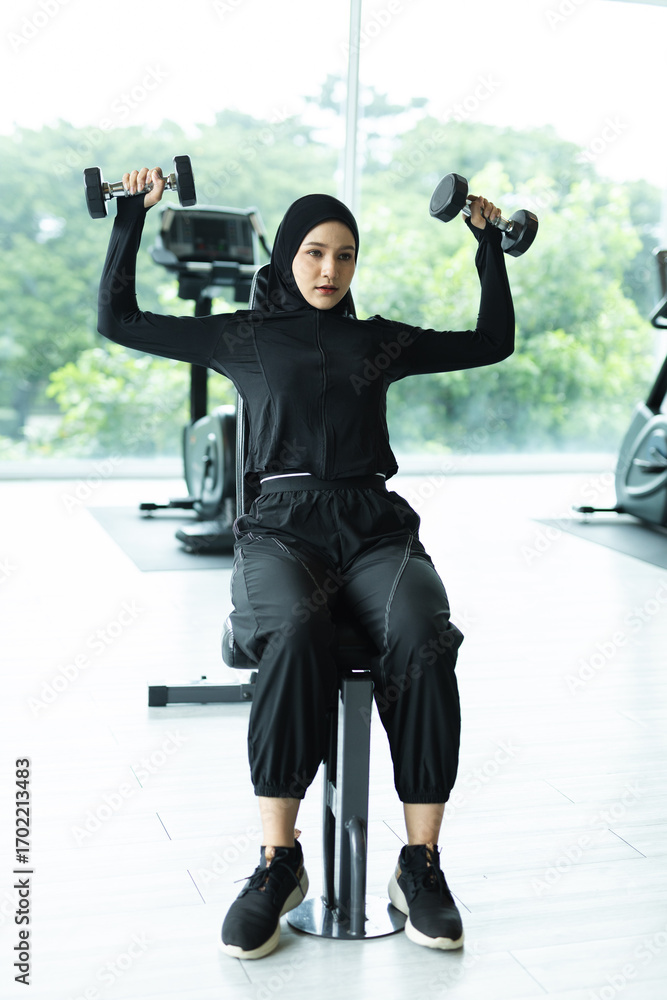 Fototapeta premium Muslim woman in sports hijab lifting dumbbells at gym, focusing on strength training and fitness, symbolizing empowerment, health, active lifestyle and diversity in modern Islamic culture.