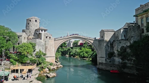 4k Still drone video of Old Bridge in Mostar, Bosnia and Herzegovina, showing tourists crossing and a speed boat passing on the Neretva River below, with the old town and mosque in the background.