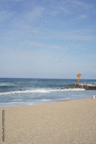 Orange lighthouse on breakwater with crashing ocean waves, sandy beach coastline and scenic seascape view under blue sky at seaside landscape