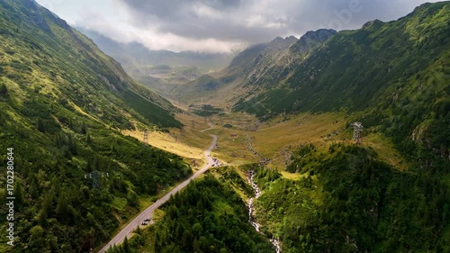 Majestic mountain valley with winding road and river. Vast green valley surrounded by mountains features a winding road and a bubbling stream under a cloudy sky.