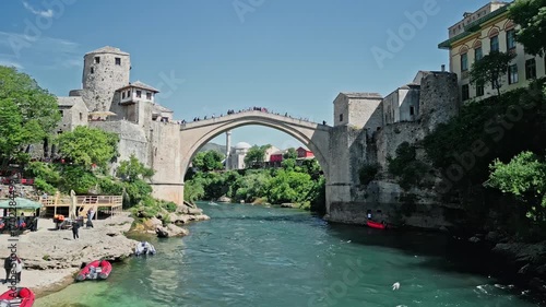 4k Dolly drone video along the Neretva River in Mostar, starting at Lučki most and flying close to the water, passing under Stari Most, ending with Koski Mehmed Pasha Mosque in view.