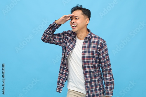 Side view of young Asian man wearing casual clothes, looking into the distance, hand gesture on forehead, isolated on blue background.