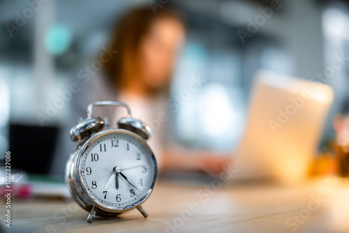 Alarm clock foreground with blurred woman working on laptop background indoors
