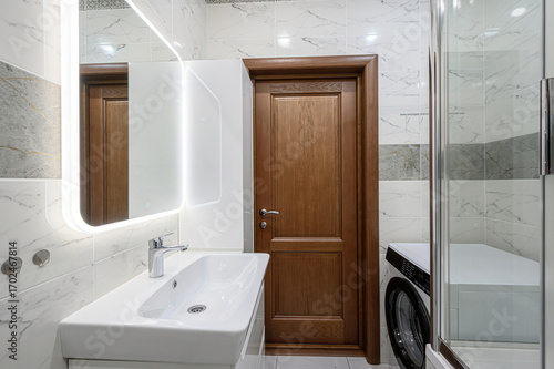 A bright bathroom features a white sink with a chrome faucet, a lit mirror, and a wood door. Marble-like tiles adorn the walls, with a glimpse of a washing machine behind a shower enclosure