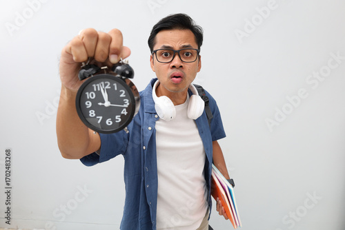 Asian male student with glasses wearing casual clothes, backpack, headphones around his neck, showing his alarm clock up while carrying a book, shocked expression looking at the camera.