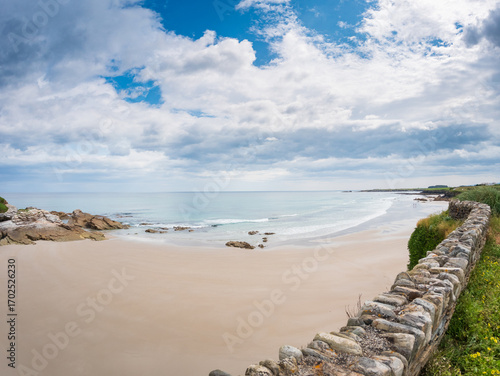 Wide view of Playa de las Catedrales in Galicia, Spain, showing sandy beach, rocky formations, calm waves, stone wall, and dramatic cloudy sky along the Atlantic coast.