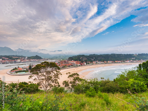 Coastal town of Ribadesella in Asturias, Spain, with sandy river mouth curving into the sea, colorful houses, mountains in background, cloudy sky above, blending culture and nature.