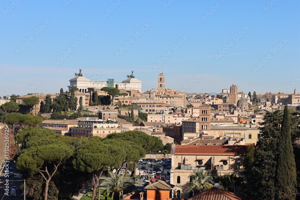 Fototapeta premium Panoramic view of Rome with historic buildings, domes, towers, and the Vittoriano monument under a clear blue sky, framed by green trees.